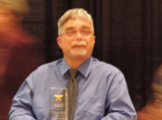Chris Mitchell wearing a blue shirt, dark colored tie, and glasses with some facial hair, sitting in a chair holding the Missouri Association of Hospital Auxiliaries Volunteer of the year award