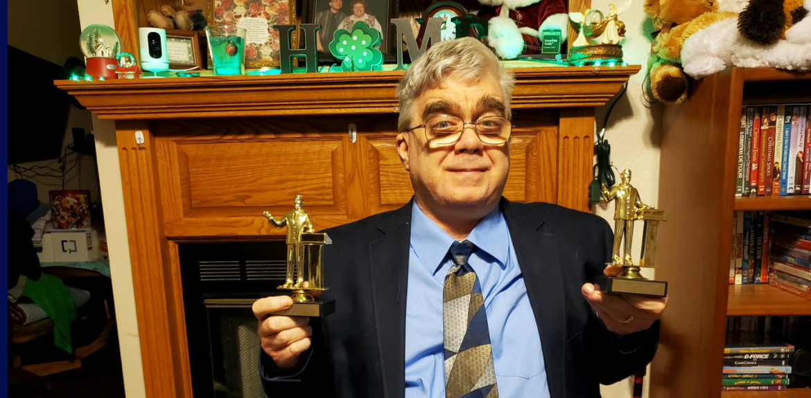 Chris in front of a fireplace with photos, security camera, green shamrocks on top next to bookcase of DVDs. Chris is holding two Toastmasters awards he earned in speech competitions. Chris is wearing glasses, short and curly hair, a light blue dress shirt, a multi-colored tie and a dark blue dress jacket