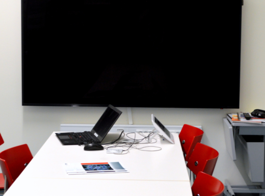 Large black monitor mounted on a white wall in a conference room. Conference table is visible with a laptop with cables attached to a tablet along with a pamplet sitting on the table. There are red chairs on each side of the table. There is a small table in the corner with a few miscellaneous items sitting onto of the table.