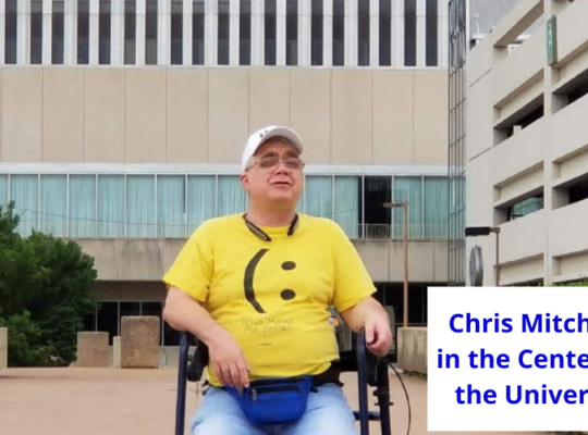 Chris Mitchell, CEO and founder of #DefineYourself, wearing a yellow happy face shirt, white ballcap, sitting on his rollator in the center of the universe in Tulsa OK