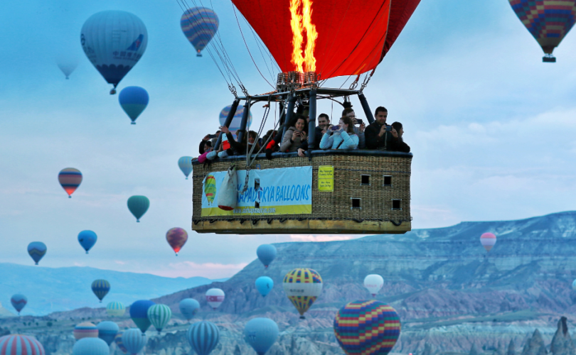 Visible is the top of a red hot air balloon flying against with several people in the basket against a blue sky and mountain background with about 3 dozen other balloons of fin the distance.