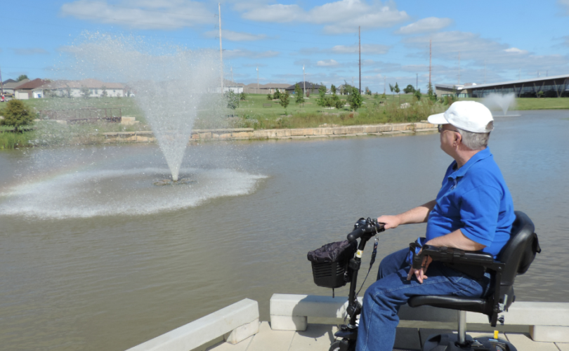 A photo of Chris siting on a black and blue scooter wearing a blue jacket and a white cap, with his back towards the camera, looking at a water fountain in a local park.