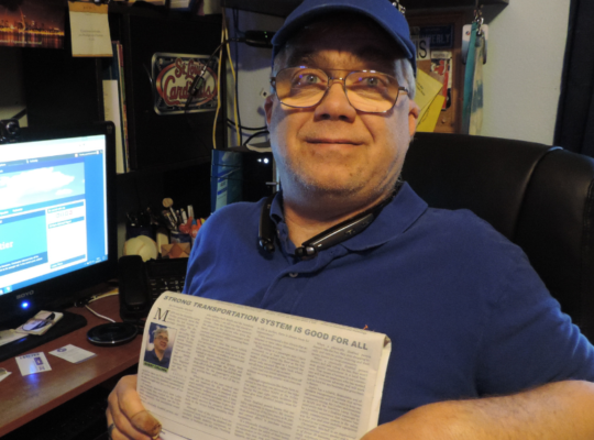 Chris Mitchell, wearing a blue ballcap and polo shirt, sitting in his office near his computer, holding up the Joplin Regional Business Journal open to his op-ed article: Strong Transportation System is Good For All
