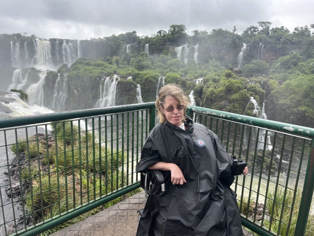 A Caucasian woman with blonde hair, wearing black sitting in an electric wheelchair with a scenic background.