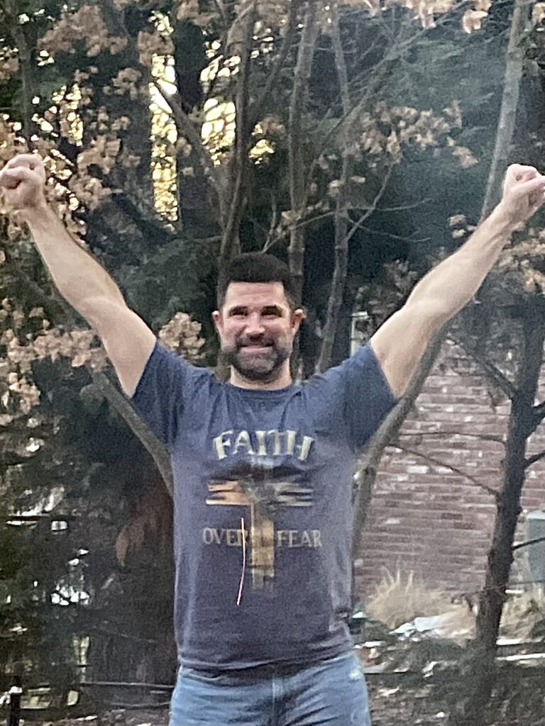 Trent Brock, a Caucasian male with dark hair and a beard, wearing a grey t shirt in the victory pose